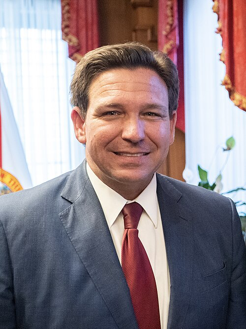 Florida Governor Ronald D. DeSantis smiling in a gray suit with red tie, photographed in his official office with decorative red curtains and state emblems visible in the background.