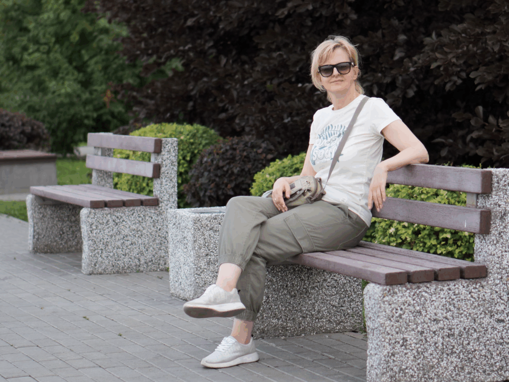 Woman with sunglasses sitting on a park bench with legs crossed while wearing pants, demonstrating why women cross their legs even when wearing comfortable clothing that doesn't require modesty considerations.