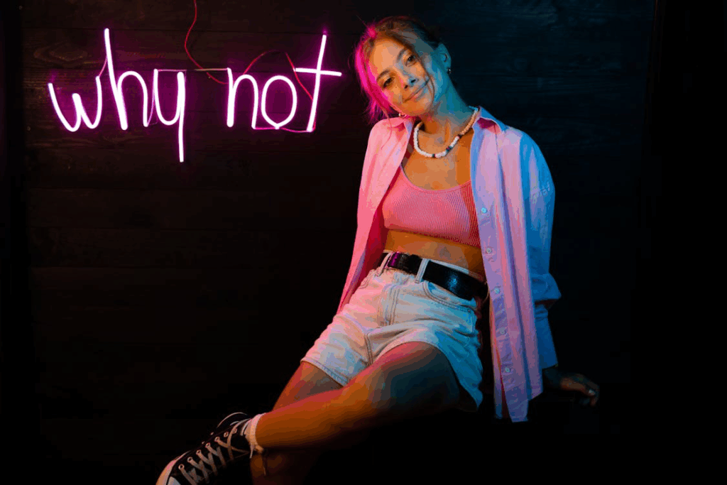 Young woman in casual clothing sitting with relaxed posture in front of neon 'why not' sign, representing the modern shift toward personal choice in sitting positions rather than following strict gender rules.