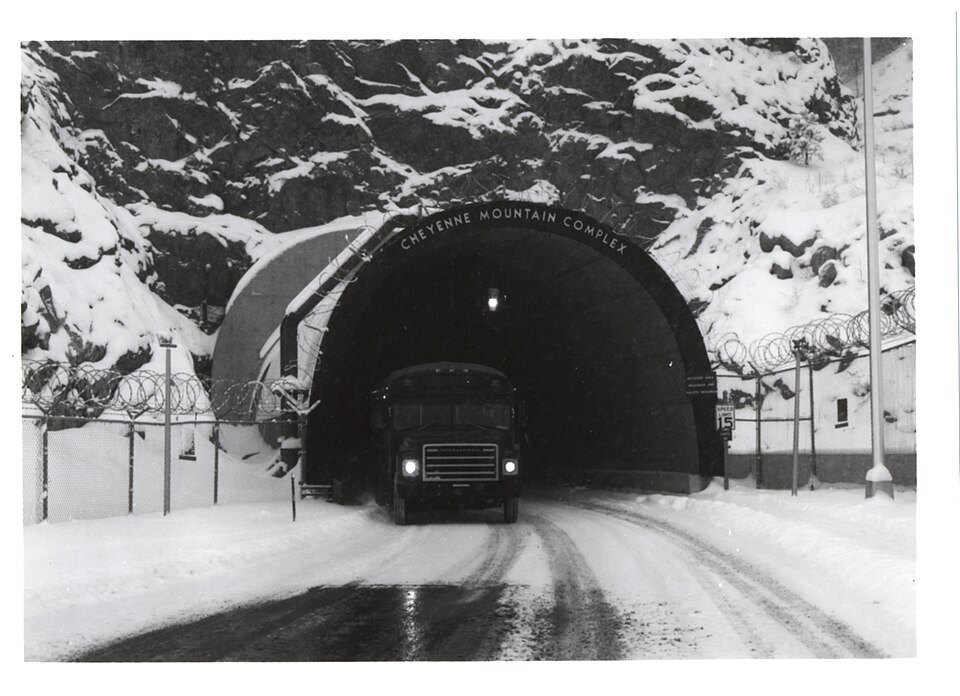 Entrance to the Cheyenne Mountain Complex in Colorado during winter, showing a snow-covered tunnel entrance with 'CHEYENNE MOUNTAIN COMPLEX' visible above the arch. A military vehicle approaches the entrance, surrounded by security fencing with barbed wire. This documented underground military facility differs from alleged secret underground cities.
