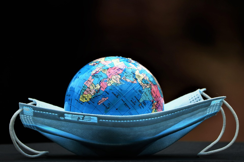 Symbolic image showing a small globe nestled inside a protective face mask against a dark background. The world map is visible through the blue medical mask, representing global health protection and the international nature of pandemic preparedness. This visual metaphor illustrates how public health measures are needed to shield the entire planet from threats like avian influenza.