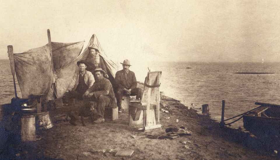 Historic photograph of four men in early settler attire seated near makeshift canvas shelters along a vast shoreline. This rare image from the Sarah A. Mooney Memorial Museum in Lemoore, CA captures one of the only surviving visual records of the original inland sea that once dominated California's Central Valley before agricultural development.
