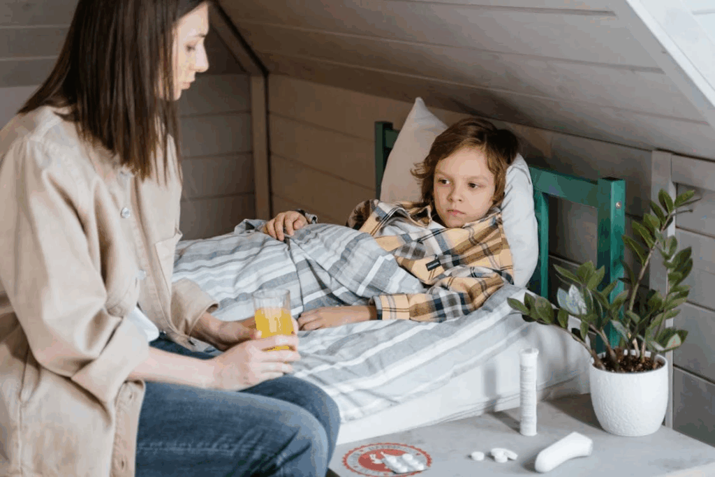 Woman sits beside child lying in bed under striped blankets with medicine nearby during whooping cough illness recovery.