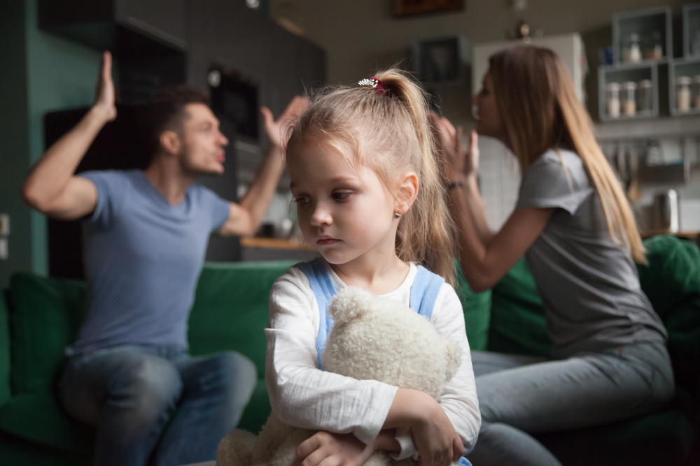 Kid daughter feels upset while parents fighting at background, sad little girl frustrated with psychological problem caused by mom and dad arguing, family conflicts or divorce impact on child concept
