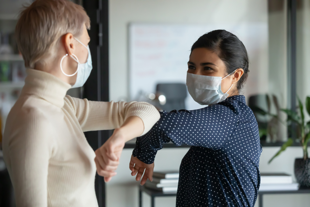 Smiling diverse female colleagues wearing protective face masks greeting bumping elbows at workplace, woman coworkers in facial covers protect from COVID-19 coronavirus in office, healthcare concept
