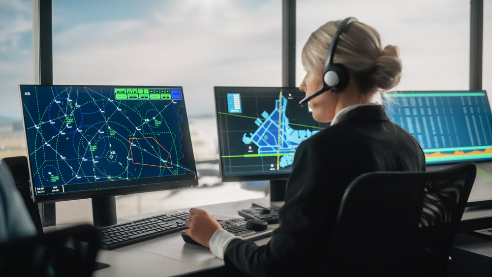Female Air Traffic Controller with Headset Talk on a Call in Airport Tower. Office Room is Full of Desktop Computer Displays with Navigation Screens, Airplane Flight Radar Data for the Team.