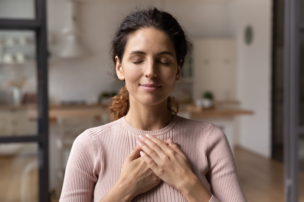 Close up of calm young Caucasian woman hold hands on heart chest feel grateful and thankful. Happy millennial female show gratitude, love and care, pray or visualize. Religion, faith concept.
