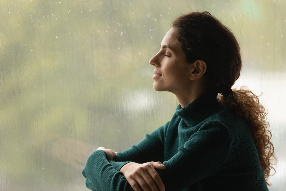 Music of rain. Side shot of serene latin woman relax by window with closed eyes listen sound of raindrops feel pleasure. Calm young lady enjoy breathing fresh ozonized air after rainstorm. Copy space.
