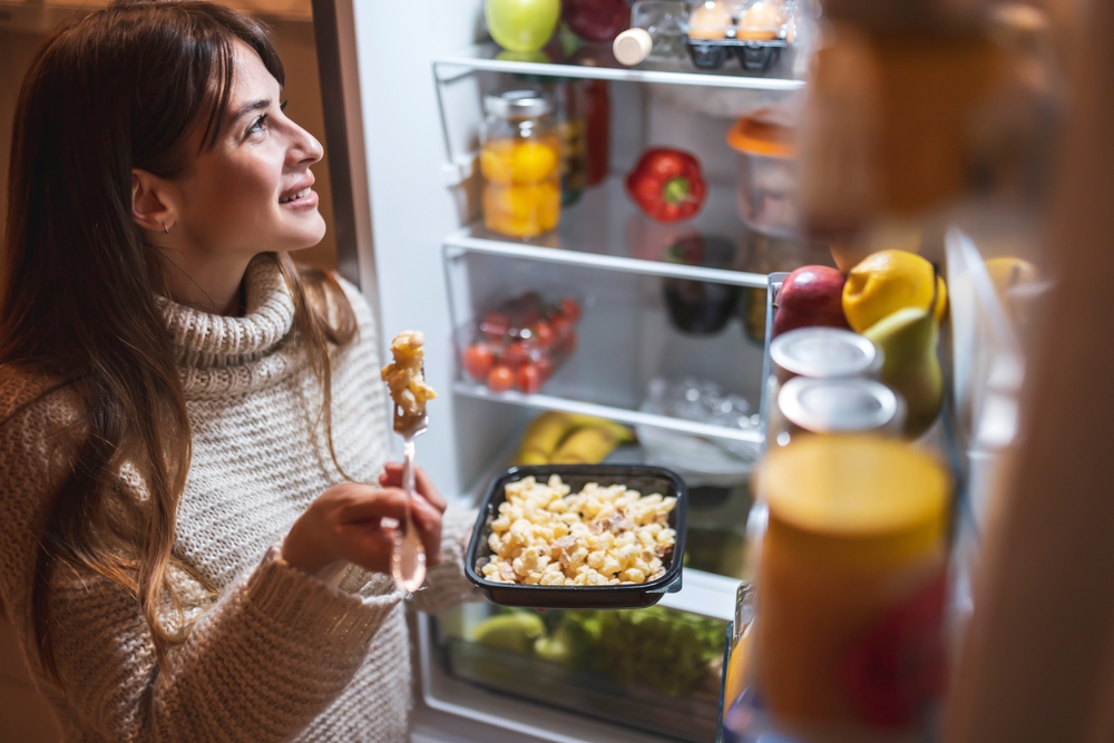 High angle view of a beautiful young woman standing next to an opened fridge door at night, eating pasta and choosing food
