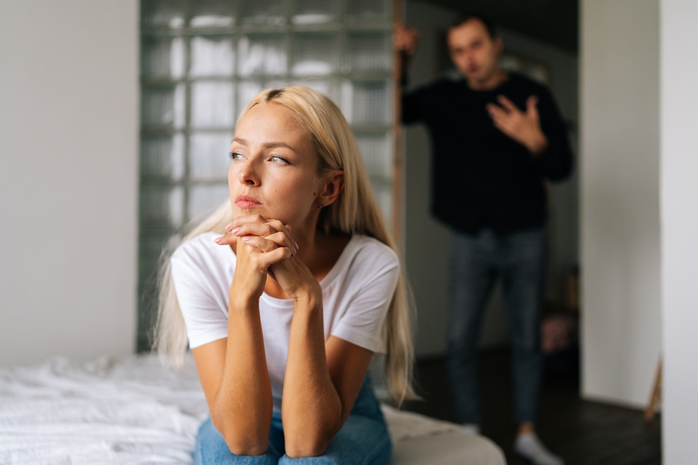 Portrait of tired stressed woman sadness looking away sitting on bed on blurred background of aggressive husband shouting on wife at home. Concept of family scandal, crisis, domestic violence, abuse.

