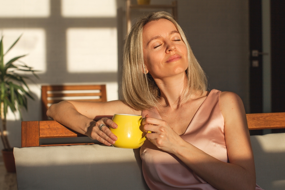 Attractive happy middle aged woman is sitting on sofa in living room. Smiling adult lady enjoys drinking coffee or tea sitting on couch at home