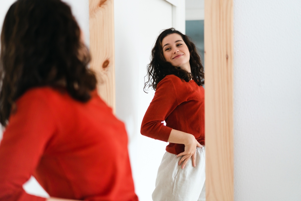 Young beautiful self confident woman in red sweater looking in the mirror proud of herself
