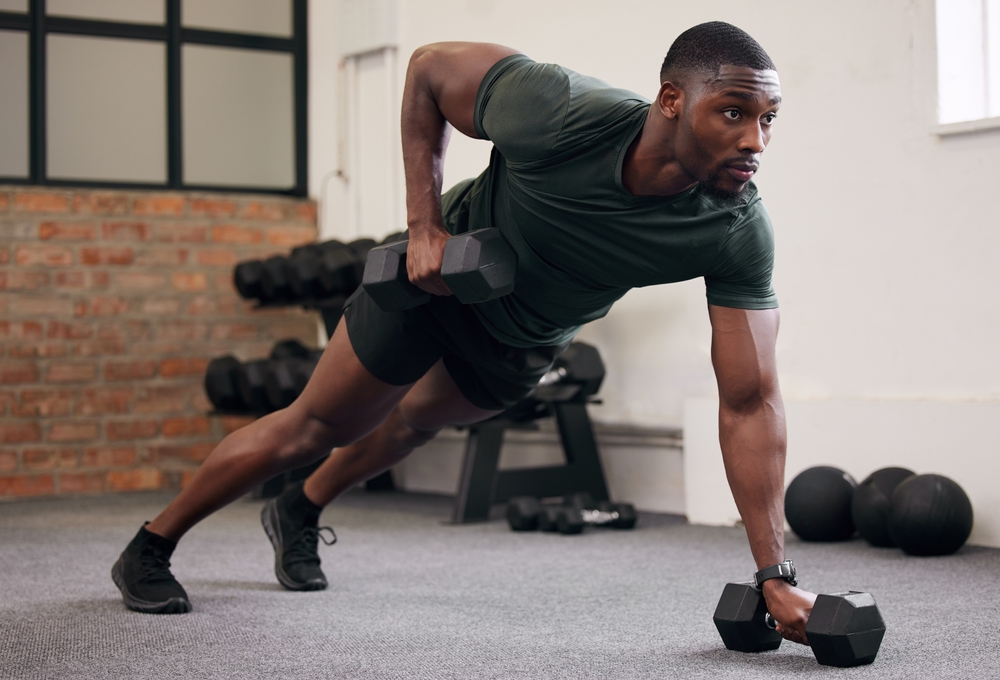Black man, dumbbell row and training in gym, workout and strong fitness in health club. Serious sports athlete, bodybuilder and weight exercise on ground for energy, power and core strength challenge
