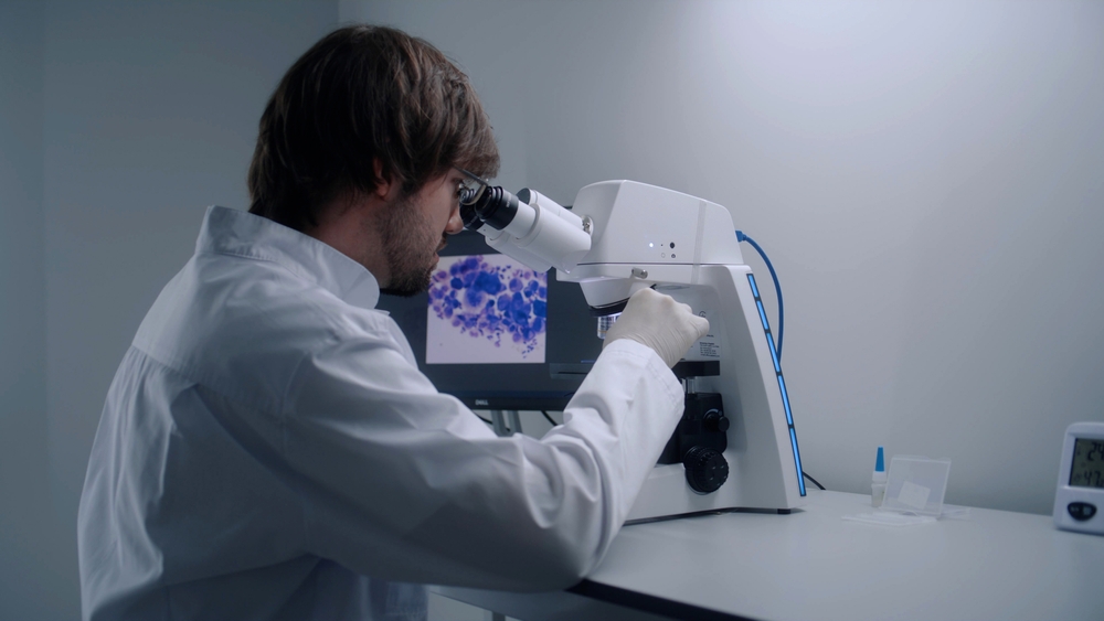 Male scientist looks under microscope, analyzes bacteria or blood sample in modern biotechnology laboratory. Work in advanced research lab. Computer screen with microorganisms image in the background.