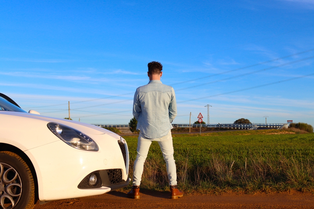 Curly haired Caucasian man peeing on a road with the car next to it. You can see the road and the sky in the background. Concept of stop to take a pee
