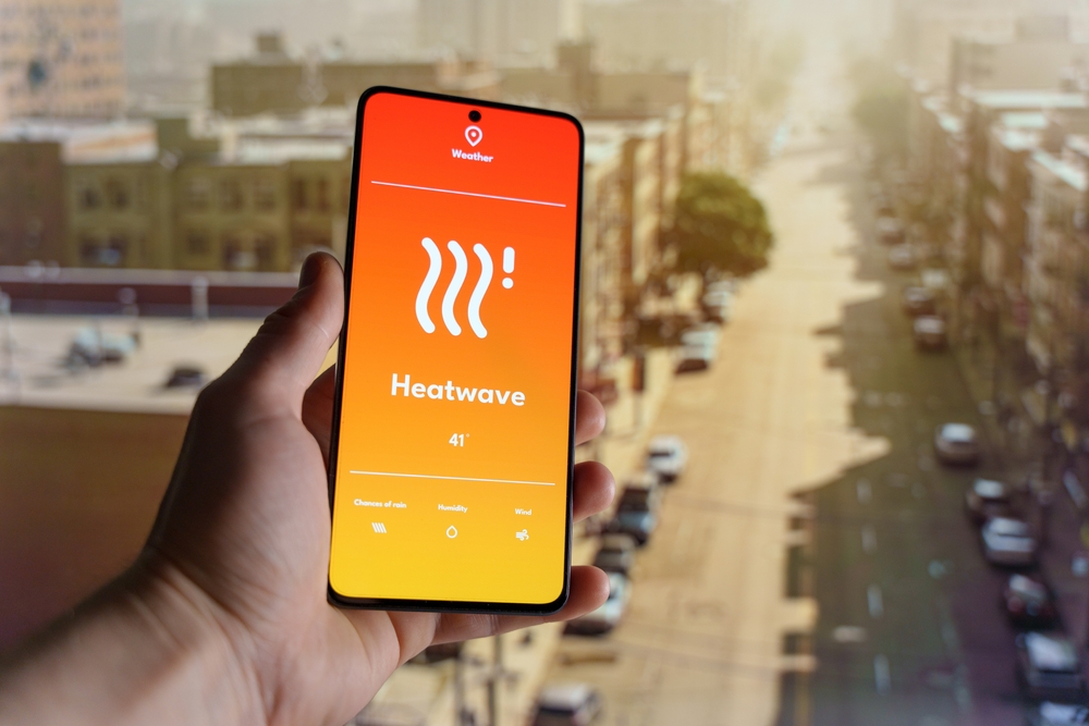 Hand holding a smartphone displaying a weather app with a heatwave warning. Blurred cityscape under extreme heat in the background. High temperature and climate change concept. High quality photo
