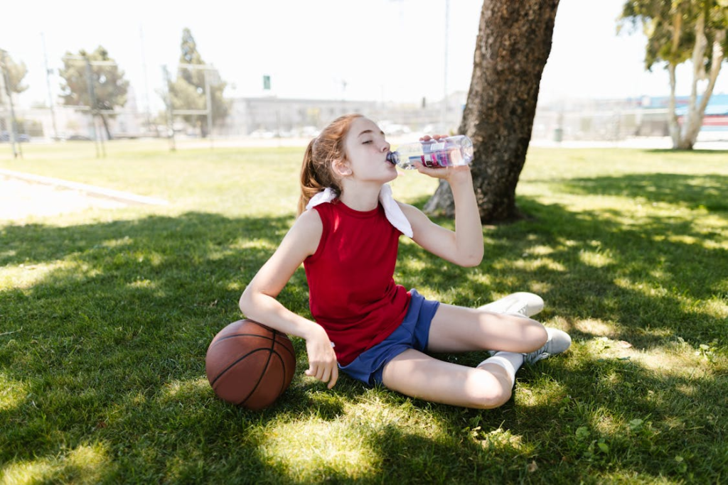 Girl in Red Tank Top and Blue Shorts Drinking Water on Grass Field
