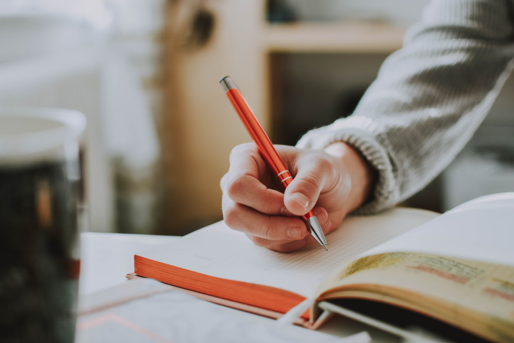 Person's hand holding red pen while writing in notebook, representing symptom tracking and documentation.