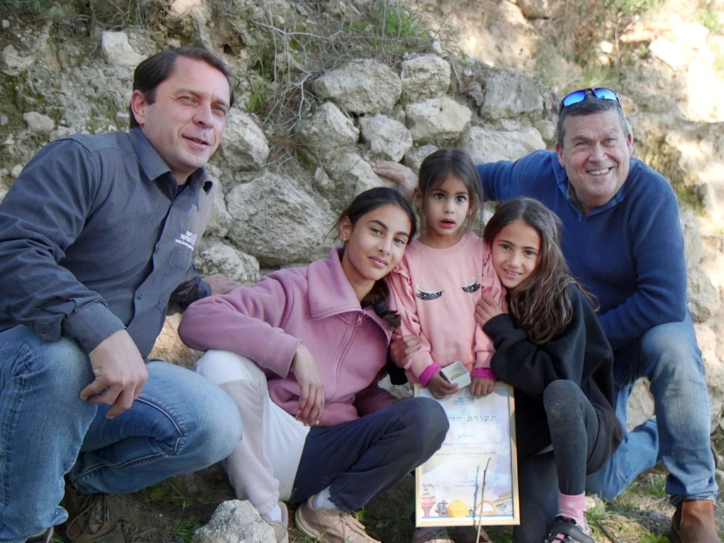 Authority)
A three-year-old Israeli girl has found a scarab-shaped Canaanite amulet dating back some 3,800 years at the site of Tel Azeka near Bet Shemesh, the Israel Antiquities Authority (IAA) announced Tuesday.
Ziv Nitzan, from Moshav Ramot Meir, was hiking with her parents and two older sisters at the foot of the hill where the archaeological site stands when she picked up what appeared to be a small stone.
“We were walking along the path, and then Ziv bent down – and out of all the stones around her, she picked up this particular stone,” Omer Nitzan, Ziv’s sister, said in a statement sent out by the IAA. “When she rubbed it and removed the sand from it, we saw something was different about it. I called my parents to come see the beautiful stone, and we realized we had discovered an archaeological find! We immediately reported this to the Israel Antiquities Authority.”
Promoted: Identity/Crisis w/ Tal Becker
Keep Watching
Scarab amulets trace their origins to ancient Egypt, where beetles were revered as sacred symbols of renewal. Egyptians believed these insects embodied new life, as they laid their eggs inside the dung balls they rolled — seemingly bringing life from decay.
“Scarabs were used in this period as seals and as amulets,” Dr. Daphna Ben-Tor, an expert in the field, said in a statement. “They were found in graves, in public buildings, and in private homes. Sometimes they bear symbols and messages that reflect religious beliefs or status.”
Get The Times of Israel's Daily Edition
by email and never miss our top stories
Newsletter email address
Your email
Get it
By signing up, you agree to the terms
Ben-Tor examined the artifact found at Tel Azeka and determined its cultural affiliation with the Canaanites, an ancient people recorded throughout the Bible and in historical documents up to 3,500 years old — as well as older archaeological findings — as a group of peoples living throughout the Levant who were ruled by their own city/state kings.
From right to left – Tel Aviv University Prof. Oded Lipschits, Noga, Ziv and Omer Nitzan, and Antiquities Authority archaeologist Semyon Gendler