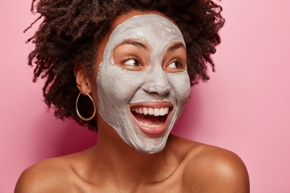 Headshot of good looking dark skinned woman with curly hairstyle, focused aside, has white clay mask on face, prepares for date with guy, does beauty treatments, isolated over pink background
