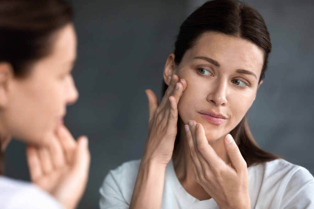 Close up unhappy sad woman looking at red acne spots on chin in mirror, upset young female dissatisfied by unhealthy skin, touching, checking dry irritated face skin, skincare and treatment concept
