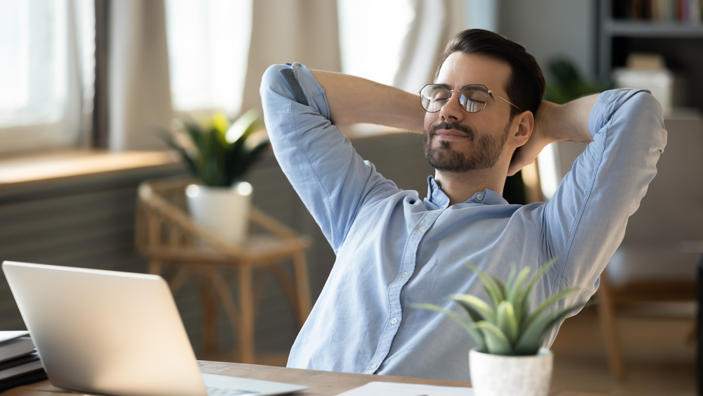 Calm millennial man in glasses sit relax at home office workplace take nap or daydream. Happy relaxed Caucasian young male rest in chair distracted from computer work, relieve negative emotions.
