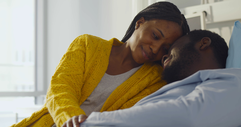 Happy african-american wife visiting smiling husband in hospital. Portrait of sick black man resting in hospital bed and embracing young woman visitor
