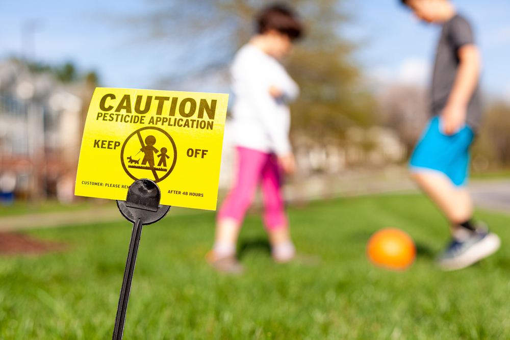 A yellow yard sign warning kids and pets of the recent pesticide spraying and advices them to stay away. Kids are playing soccer in the background regardless. Pesticide use is a big concern.
