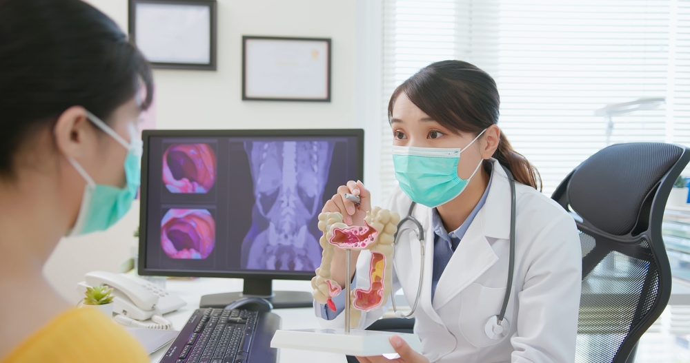 asian woman wears face mask to prevent COVID19 has colorectal cancer diagnosis in hospital - female doctor showing colonoscopy results and xray to patient on computer and explain by anatomical model
