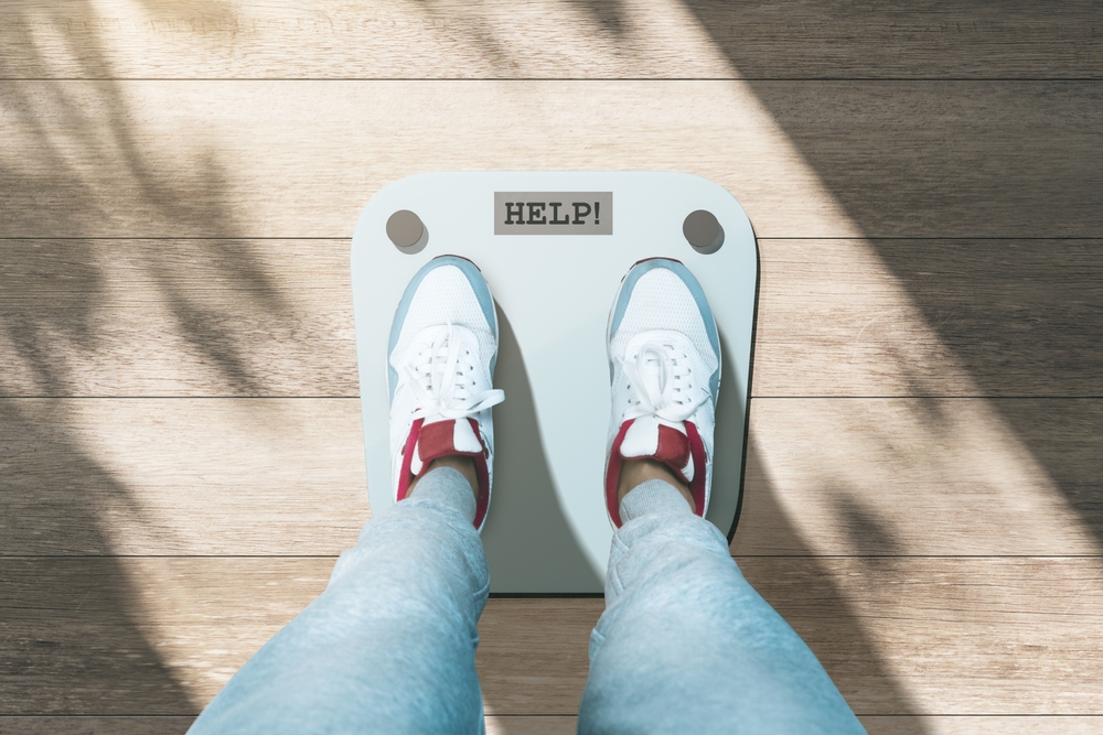 Close up and top view of creative casual dressed feet on scales asking for help. Lose weight and measuring concept. Wooden floor background with shadows