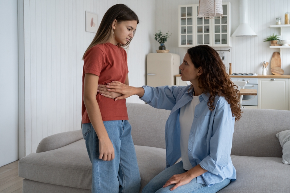 Stomachaches in Kids. Unwell pale teen girl daughter standing near mother complaining of stomach ache, unhealthy child kid feeling discomfort in belly area showing mom where it hurts. Parental care
