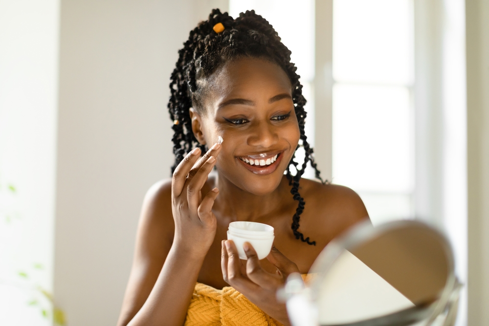 Attractive black woman using facial cream after shower, making daily skin care routine, sitting in bedroom at home, looking at mirror and smiling, free space
