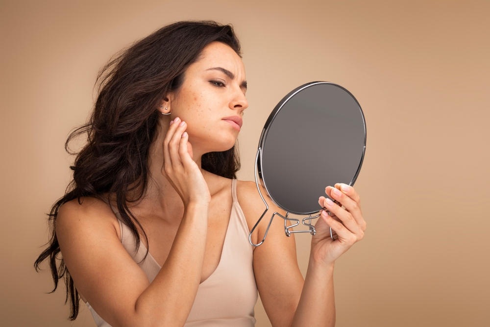 Stressed sad brunette young woman wearing beige top looking at mirror and touching her face over beige studio background, having skin problems. Acne, pimples, wrinkles, dull skin
