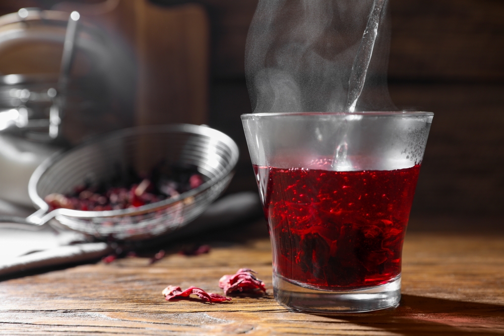Making hibiscus tea. Pouring hot water into cup with roselle petals at wooden table, closeup. Space for text
