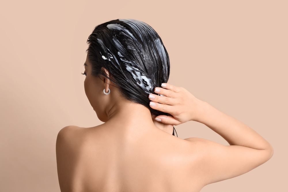 Young woman applying hair mask on beige background, back view
