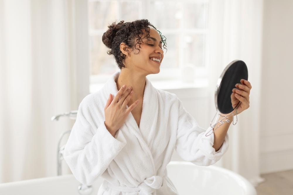Happy beautiful black woman in bathroom and looking at mirror, checking her neck, enjoying results after massage or home spa treatment
