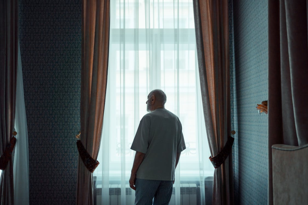 Old Man Standing by the Window Looking Outside
