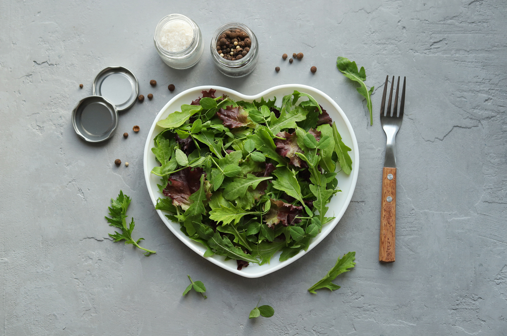 Fresh salad mixture of arugula leaves, mizuna lettuce, peas and lollo rosso in a heart-shaped plate, salt and spices on a gray concrete background. Concept: healthy food, top view.

