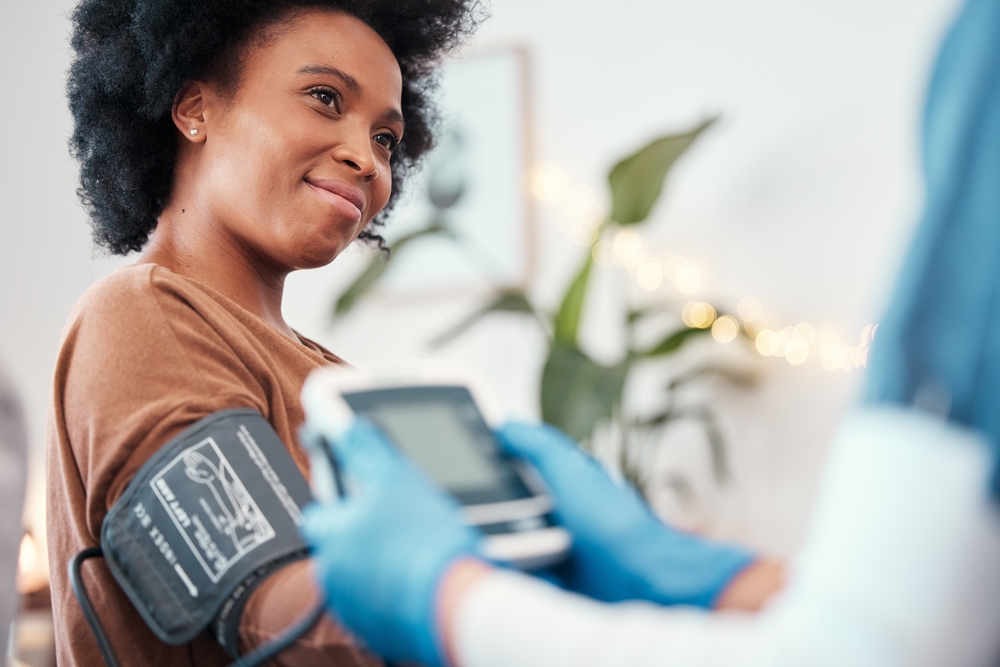 Black woman, healthcare and blood pressure machine in checkup with caregiver for monitoring pulse at home. Happy African American female patient or visit from medical nurse for health and wellness

