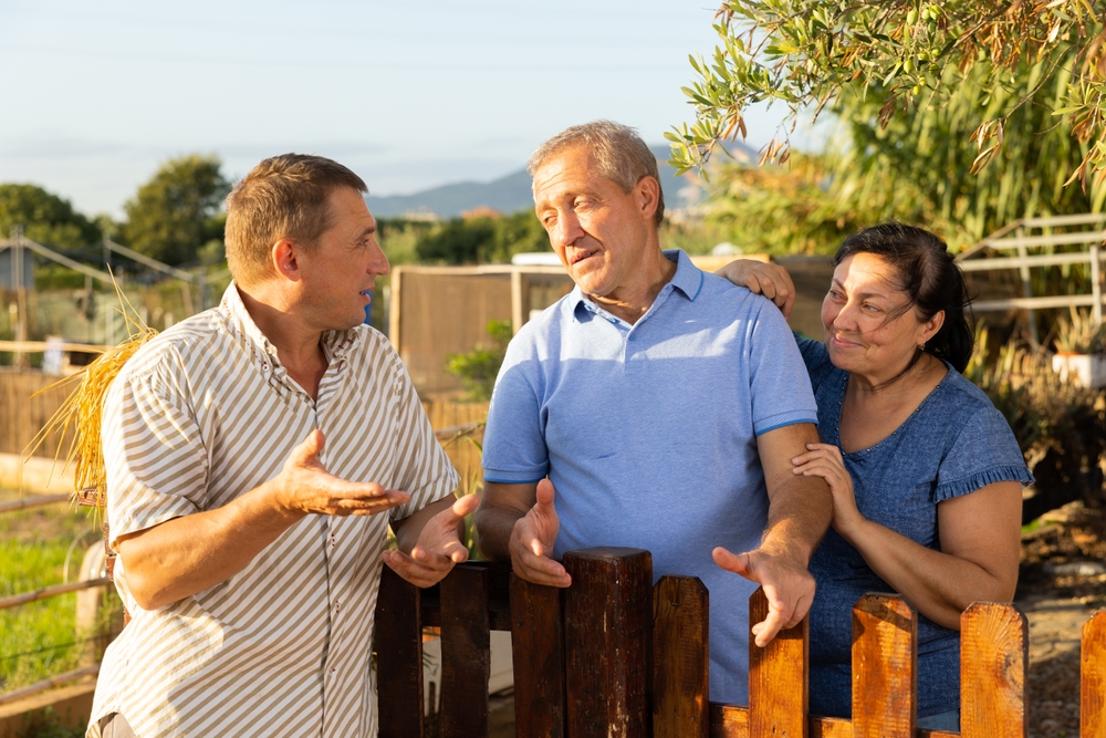 Man standing at fence and talking with mature couple neighbours at backyard garden.
