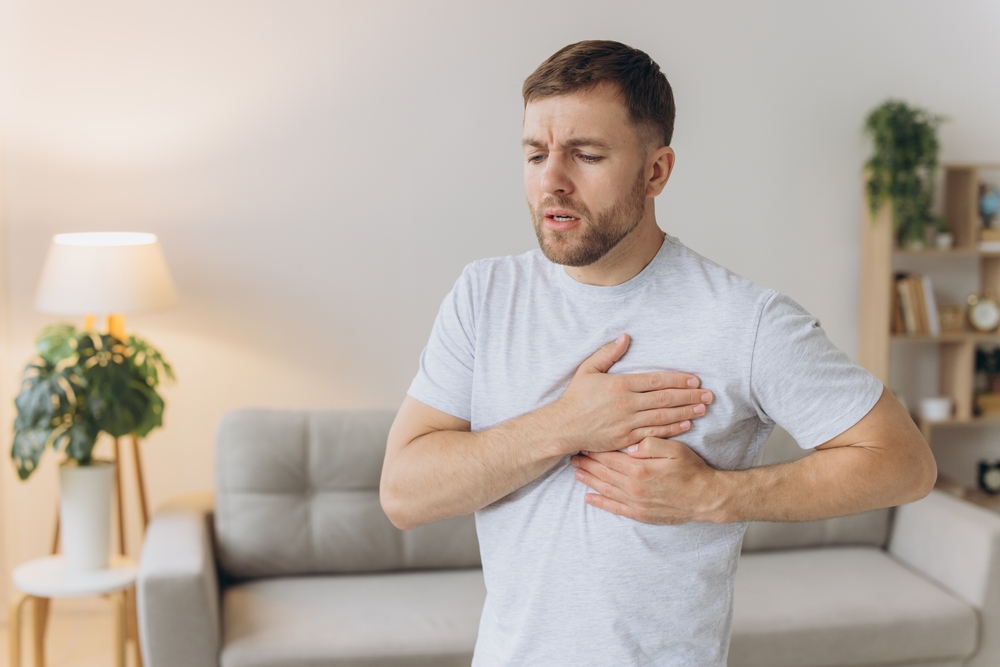 A young man is standing at home and holding his chest. Feels severe pain, shortness of breath, panic attack, heart attack, stroke.

