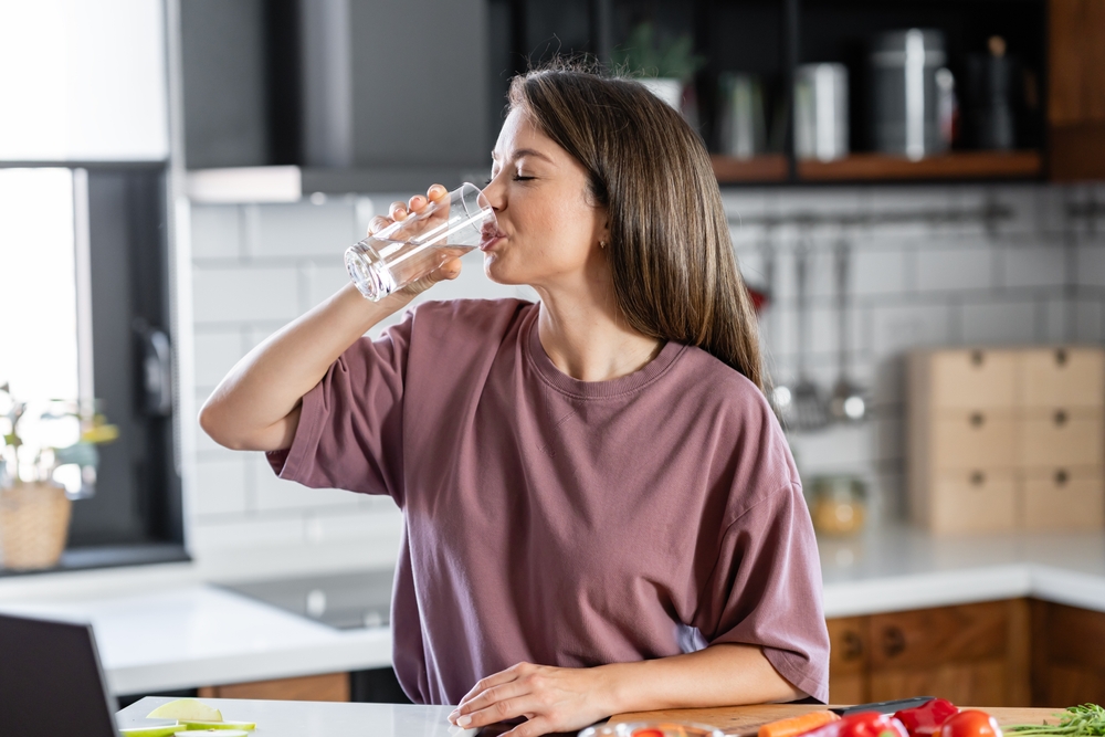 A young pretty cheerful woman is drinking a glass of water in home kitchen in the morning
