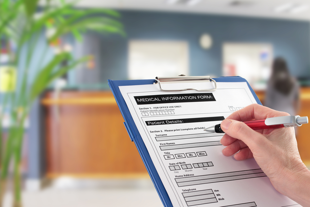 A Hand filling in medical information form with blurred hospital reception area in background
