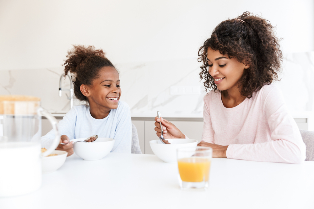 Image of african american woman and her little daughter having breakfast and eating corn flakes in kitchen