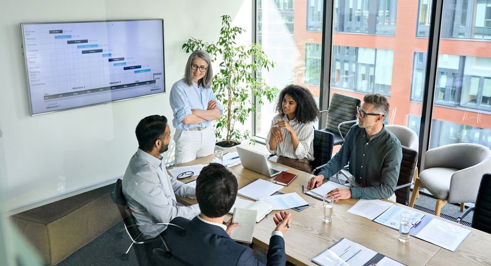 Senior female ceo and multicultural business people discussing company presentation at boardroom table. Diverse corporate team working together in modern meeting room office. Top view through glass
