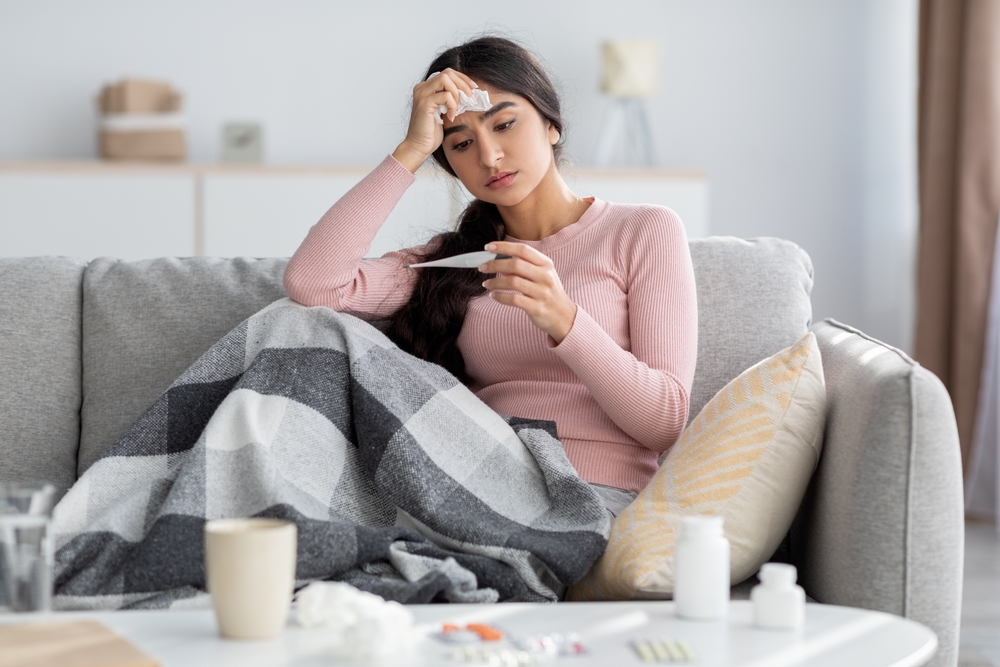 Despaired sad young hindu lady with plaid, suffer from flu, sits on sofa and looks at thermometer in living room interior. Cold, fever, disease treatment at home and covid-19 lockdown, free space