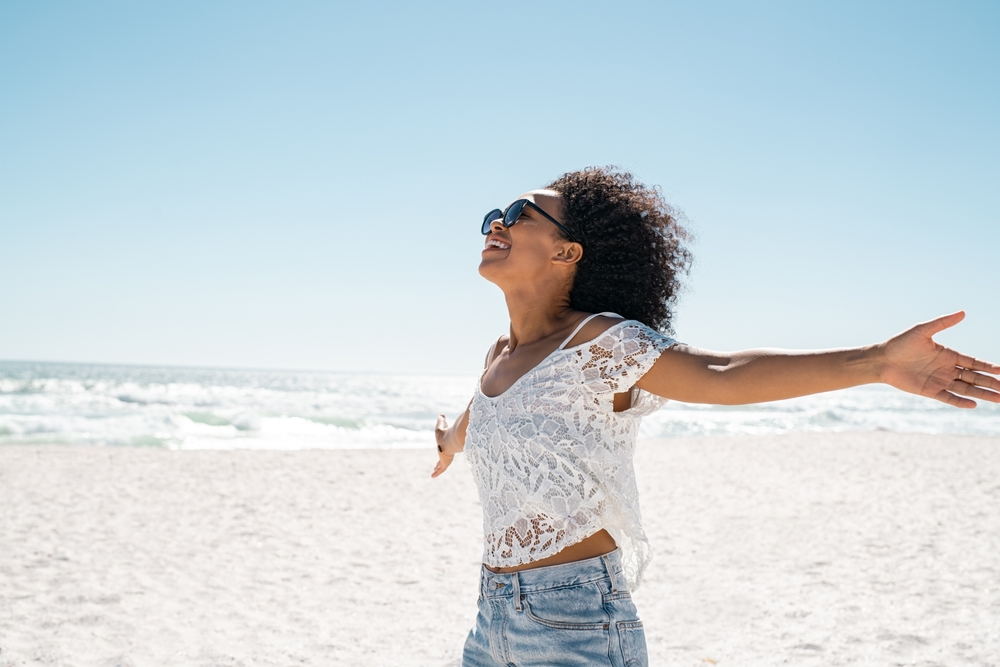 Healthy black woman standing on the beach with copy space. Happy young african american woman with open arms at seaside. Freedom girl dancing and daydreaming at beach during summer vacation.
