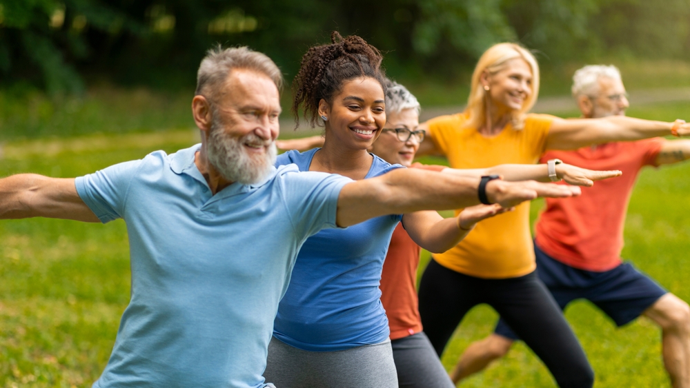 Group of diverse seniors led by young instructor practicing warrior yoga pose outdoors, happy elderly people promoting inclusivity and wellness, exercising in lush park setting, closeup shot
