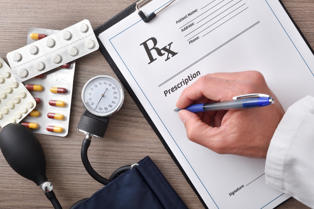 Doctor prescribing medications on a document for blood pressure on a wooden office table with blood pressure monitor and medications. Top view.