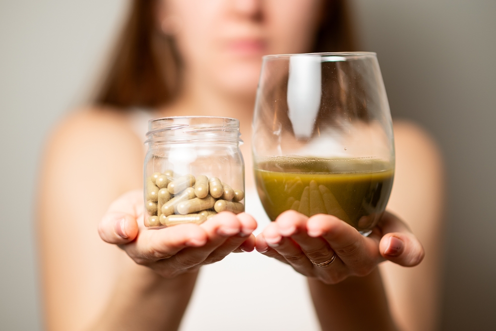 Kratom consumption concept:  female holds a ready to drink glass of green liquid, powdered kratom melted in hot water, glass of tea and handful of pills in the jar, close up photo, selective focus